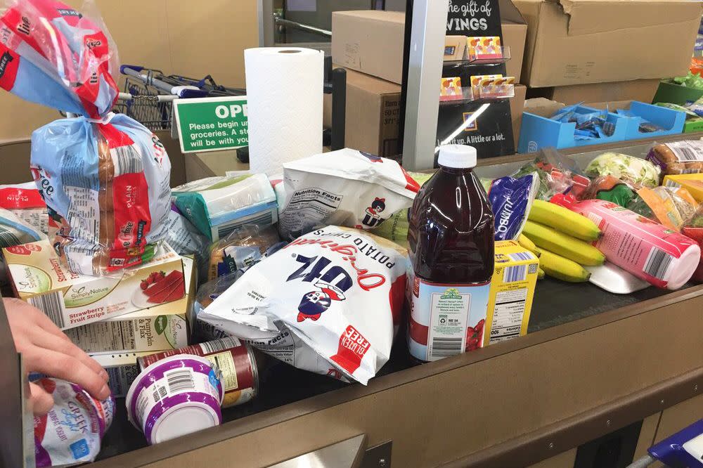 Several groceries on the checkout conveyor belt in an aldi store, an array of different kinds including potato chips, a loaf of bread, and bananas, hand on the left side putting Greek yogurt on it, with checkout items in the background like a rack of gift