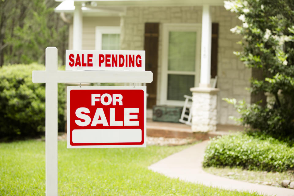 Red and white "Sale Pending, Home for Sale" sign in front of a stone, wood house that is for sale and has recently been sold. Green grass and bushes indicate the spring or summer season. Front porch and windows in background. Real estate signs in residential neighborhood.  Moving house, relocation concepts.