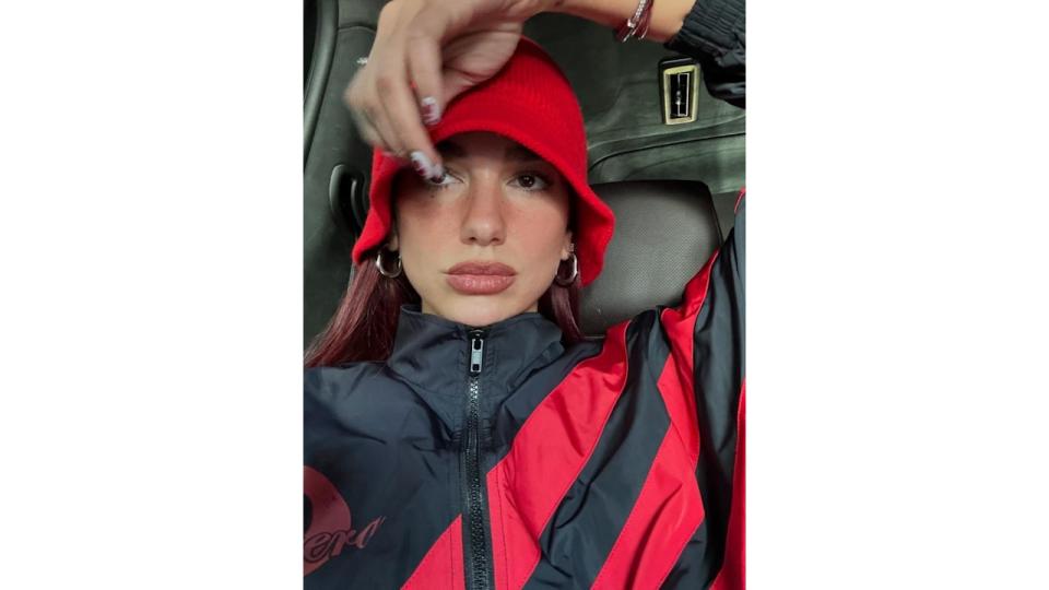 Dua Lipa poses in a car wearing a black and red jacket, a red hat and silver hoop earrings