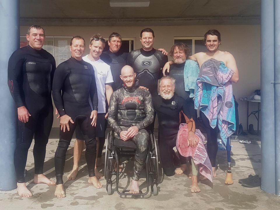 A photograph of a group of people in wetsuits smiling and embracing, dripping with ocean water.