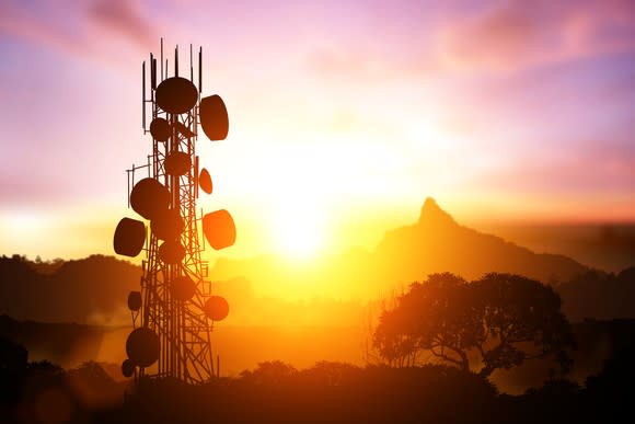 A cell tower with many signal modules in silhouette against a colorful sunrise.