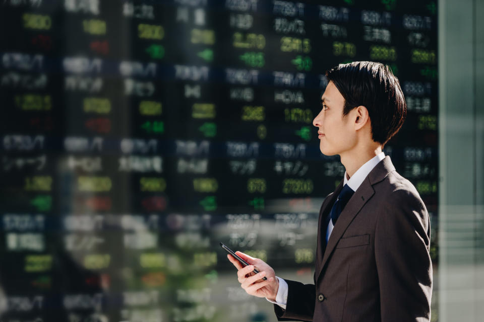 The profile of a man in a 30 -year -old suit who trades stocks with a smartphone in front of the real -time stock price board on the electric bulletin board