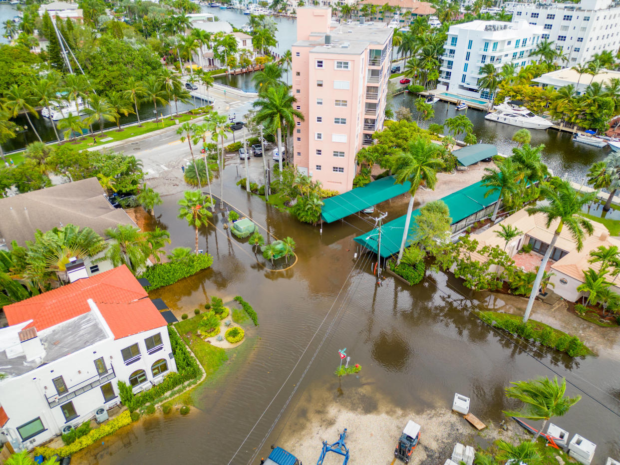 An aerial view of an eight-story building and other structures surrounded by palm trees, all marooned in water.