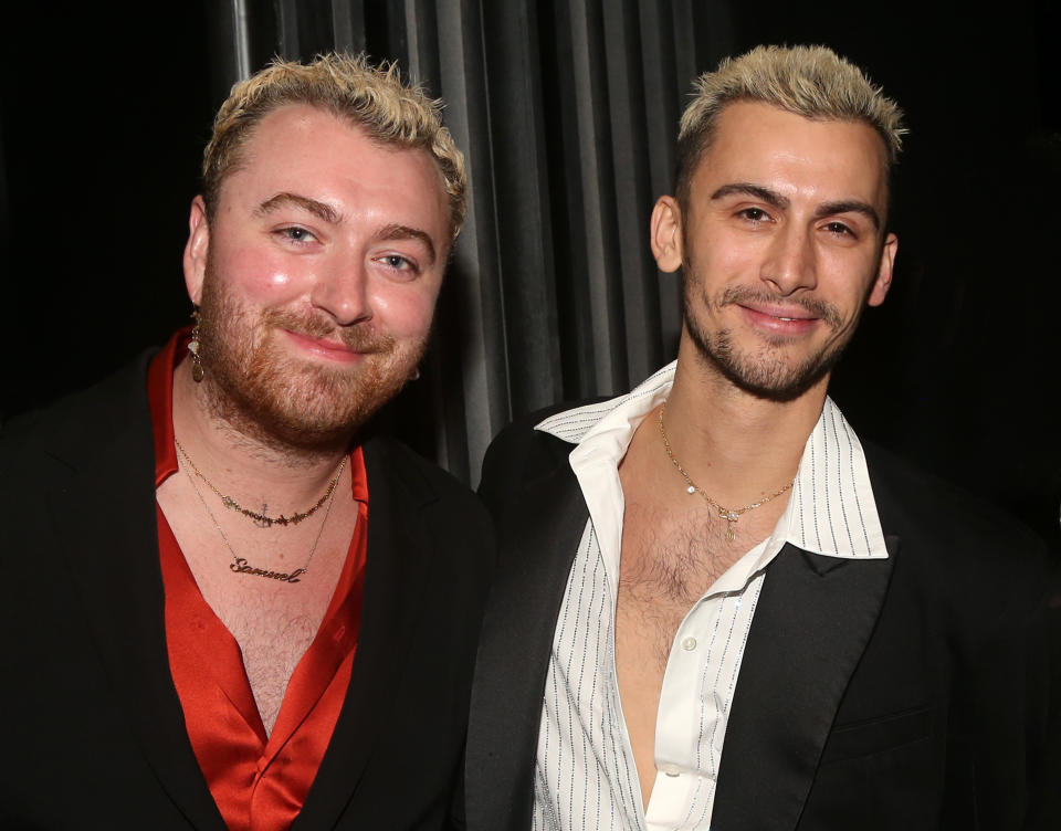 Sam Smith and Christian Cowan, both with short blond hair, smiling and posing together at an event. Sam wears a red shirt under a black jacket