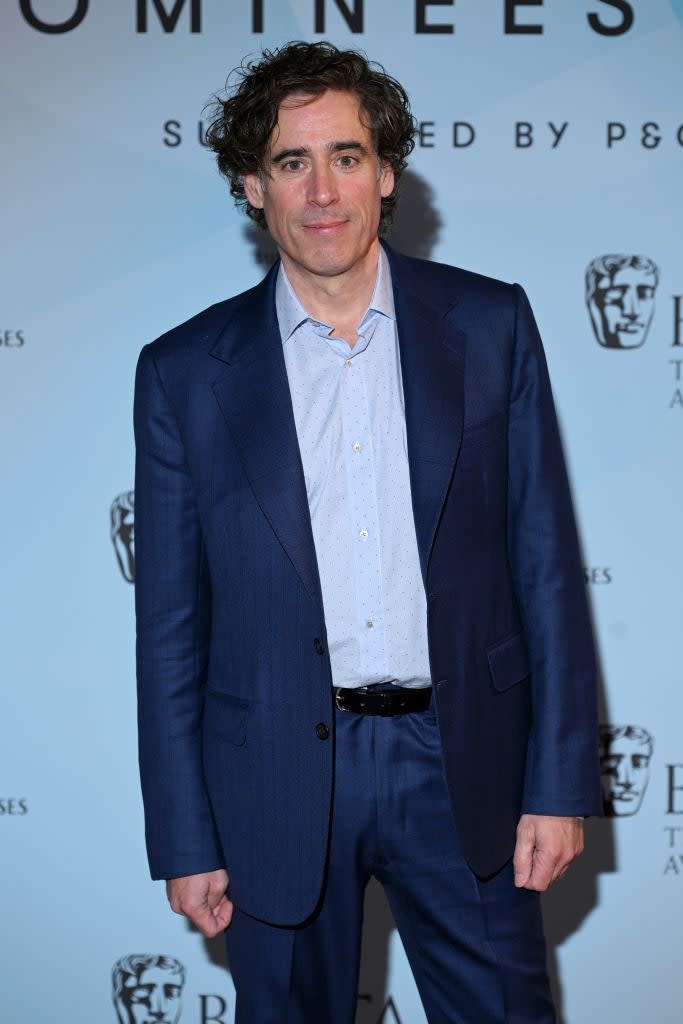 Stephen Mangan on the red carpet wearing a dark suit and a light shirt, standing in front of a backdrop with logos and text