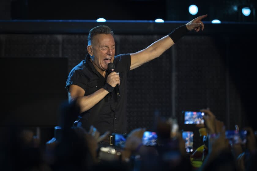 Bruce Springsteen is singing into a microphone with his eyes closed and hand raised in front of a crowd with phones out.