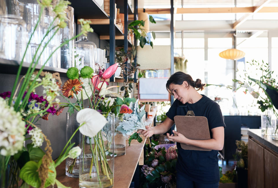 Young female florist talking on the phone with a customer and carrying a clipboard while working in her flower shop