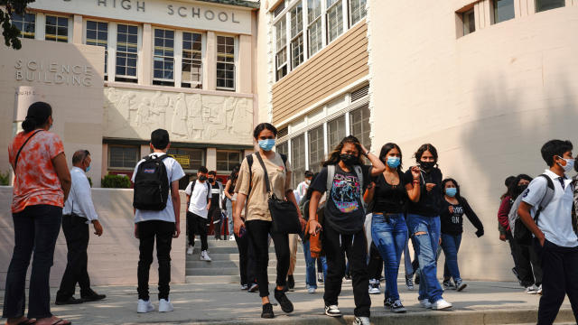 Students wearing face masks walk in a courtyard near the front of a high school building.