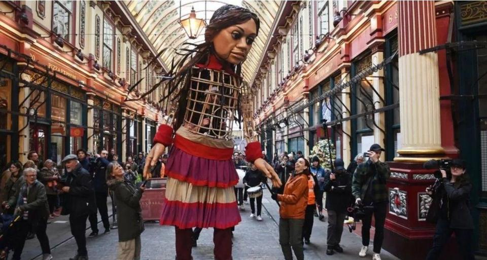 A 3.7 meter tall doll of a girl stands in an old shopping center in London as people look on 