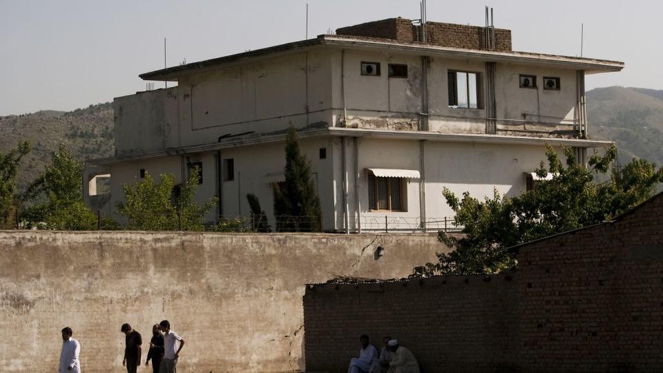 a square white house sits above a concrete wall with barbed wire, treetops appear above the concrete wall, people walk and sit outside the wall