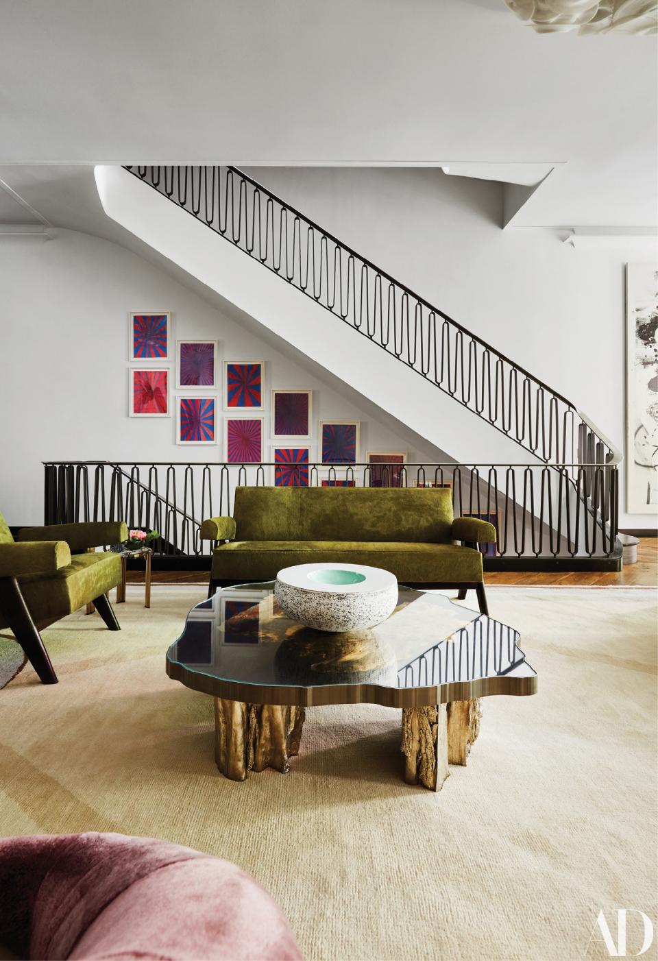 In the living room, a settee and a pair of chairs by Pierre Jeanneret surround a cocktail table by Maria Pergay. Artwork by Mark Grotjahn lines the staircase.