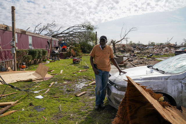 A man stands next to a car looking at the damage around him