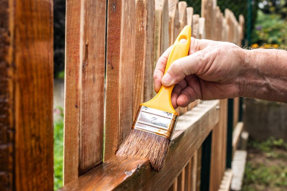 Man painting a picket fence with wood stain using a yellow-handled paintbrush.