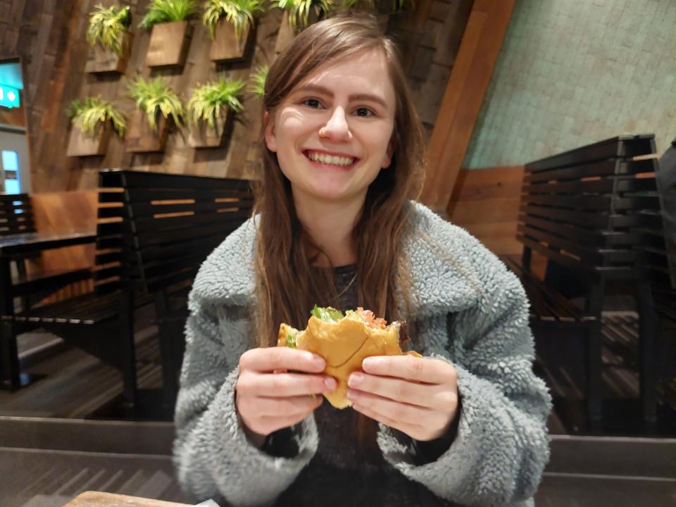 Grace Dean smiling while holding a Shake Shack burger that has been bitten into