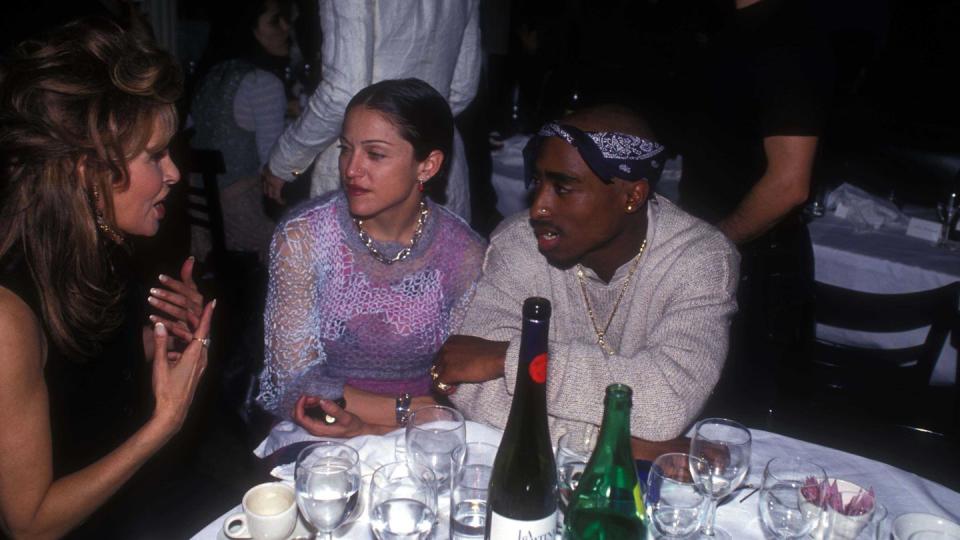 tupac shakur, wearing a white sweater and blue bandana, and madonna, wearing a pink see through shirt, sit at a table with several bottles of alcohol and glasses of water, speaking with raquel welch, who wears a black sleeveless shirt