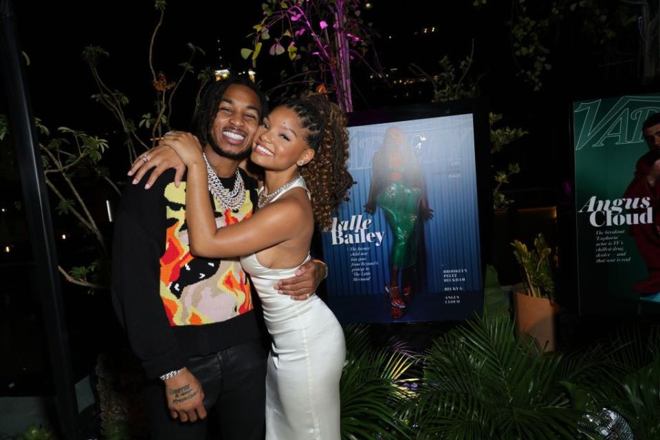 ddg and halle bailey hug and smile for a photo, he wears a graphic sweater with jeans, she wears a white halter top dress