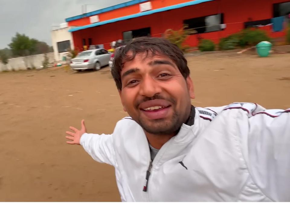 Raway smiles as he vlogs himself gesturing behind him towards a red building.