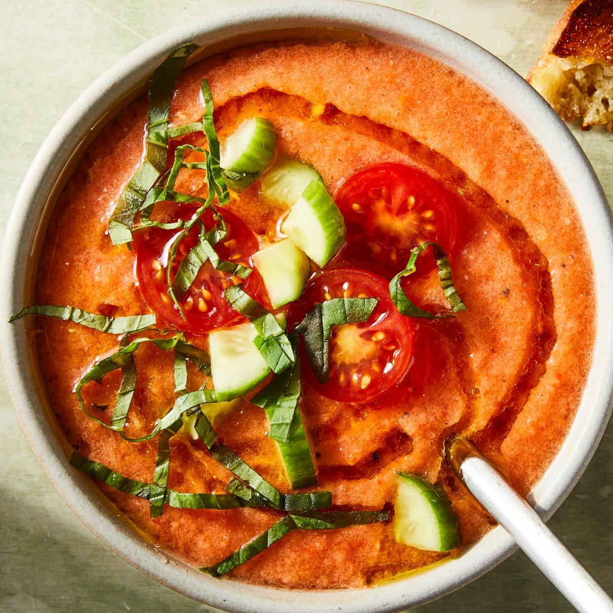 tomato gazpacho in a bowl topped with chopped cucumbers, basil, sliced cherry tomatoes, and an olive oil drizzle