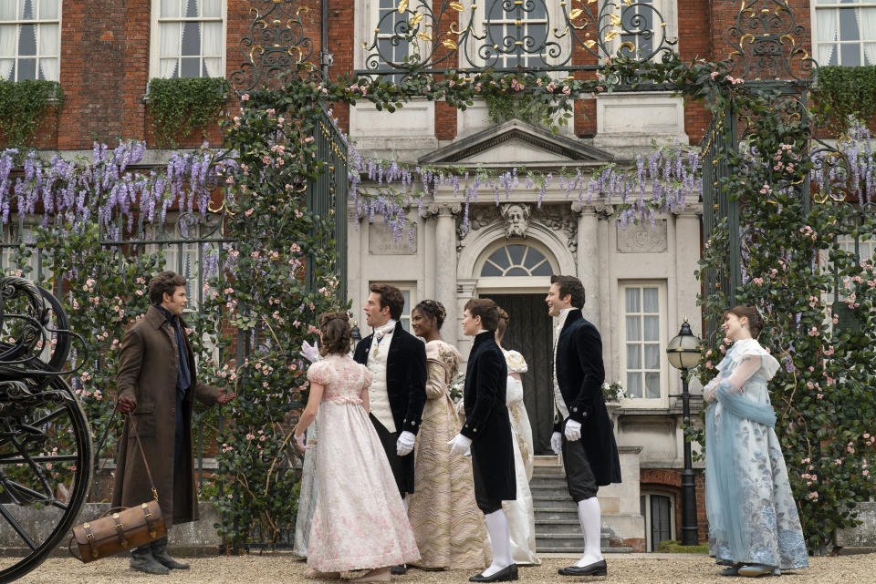 The cast of "Bridgerton" dressed in Regency-era attire, with men in black tailcoats and women in elegant gowns, converse in front of a grand estate with floral decorations