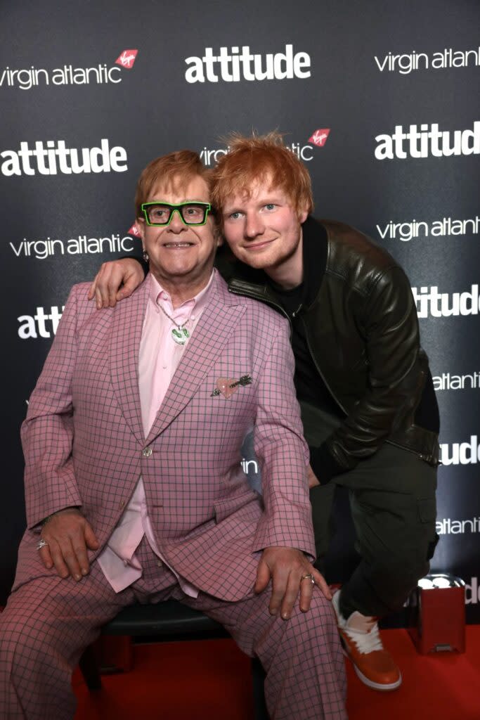 Elton John sitting down in a pink suit, with Ed Sheeran, in black, standing next to him