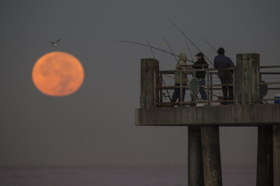 The moon sets behind people fishing on a pier in Redondo Beach, California.