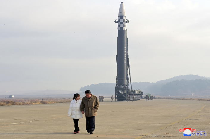 Kim Jong Un and his daughter walk across a flat military field, with an intercontinental ballistic missile towering in the background.