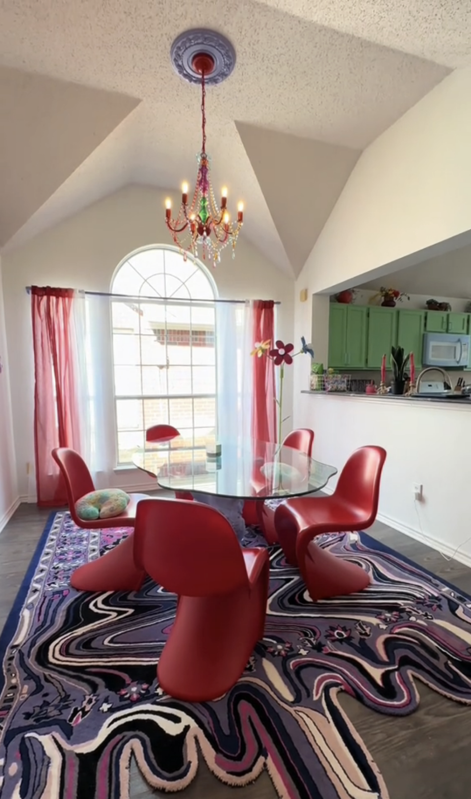 Interior room with a modern design featuring a glass table, red chairs, a colorful rug, and a chandelier