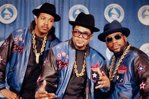 RUN-DMC poses in full regalia that they popularized- heavy gold chains, hats, sunglasses, and Adidas track suits.