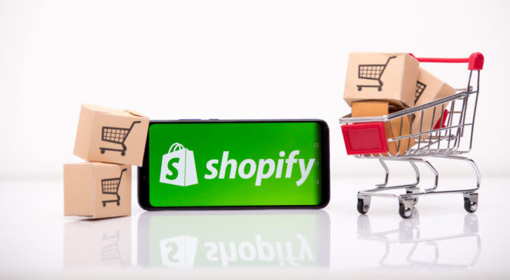 Shopify (SHOP) logo on a smartphone which is next to a miniature shopping cart and miniature cardboard boxes