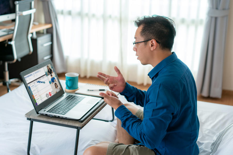 Mixed race businessman wearing business blue shirt on top and short sweatpants on bottom, showing other people on screen in formal fridays business outfits meeting online in bedroom at home.