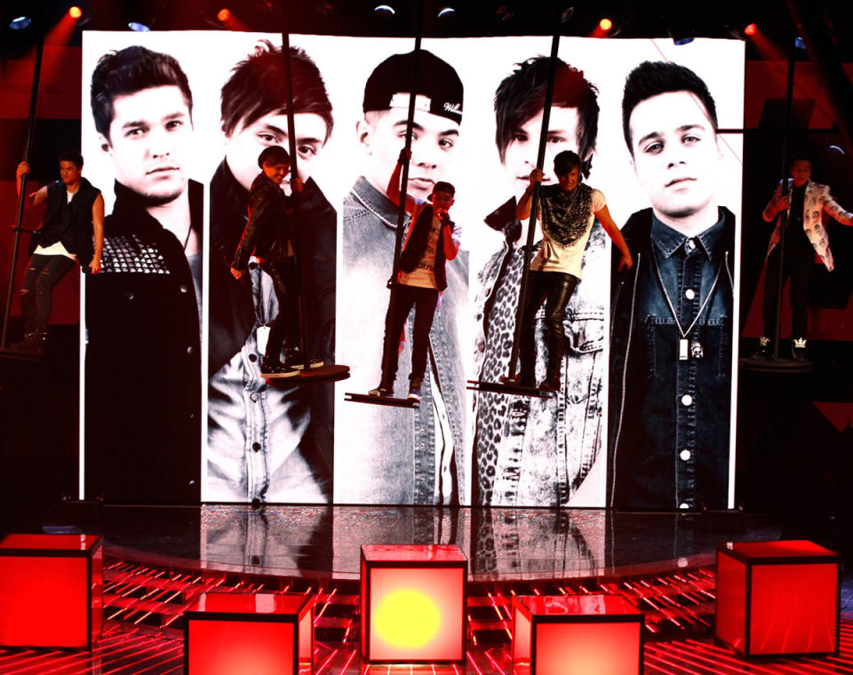 The Collective boy band are on The X Factor stage in the air, performing. There is a giant photo of the 5 boys behind them in the background.