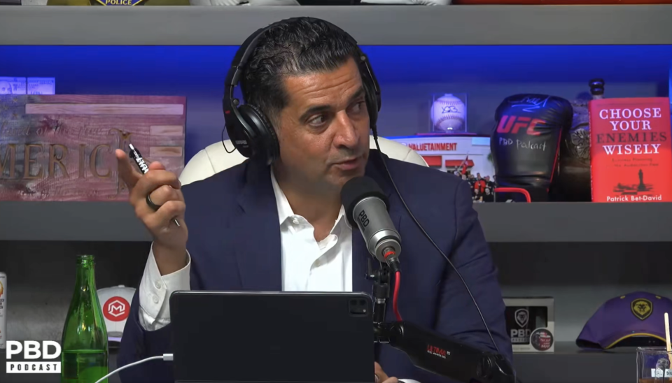 Man at podcast desk with microphone and laptop, various memorabilia on the shelf behind him