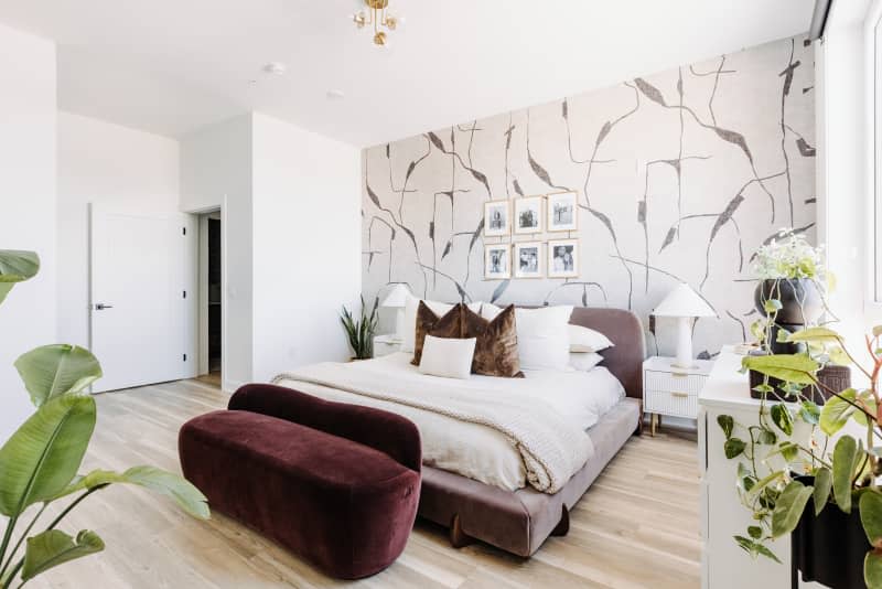 White bedroom with one gray and white patterned wallpaper wall, gray velvet bed, and other velvet accents