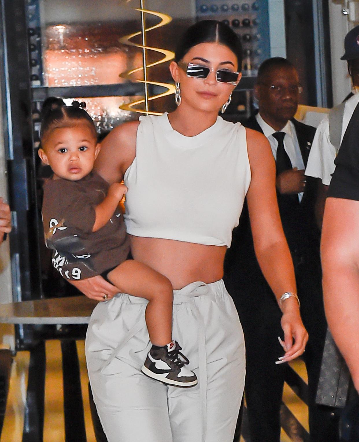 Kylie Jenner wears sunglasses with a white crop top and matching pants holding her daughter Stormi