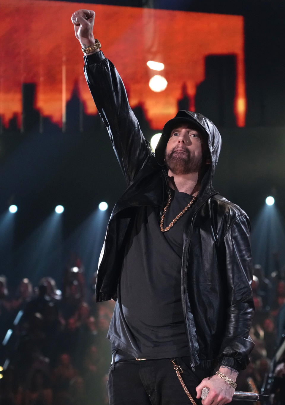 Eminem on stage in a black hoodie and jacket, raising his right fist, with a cityscape background and an audience behind him