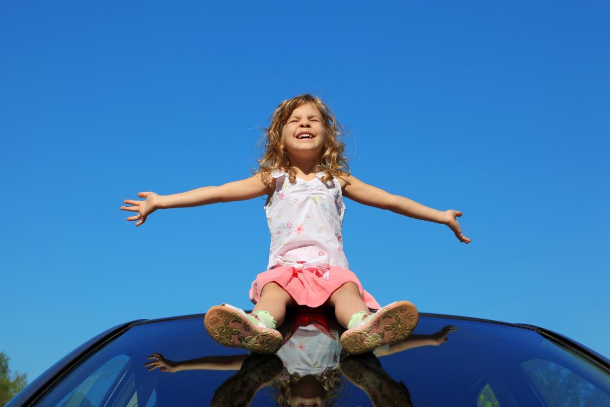 little girl sitting on car roof with open hands on blue sky