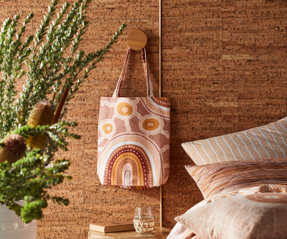 A patterned orange, red and pink tote bag in an indigenous print hangs on a brown wall next to a bed. 