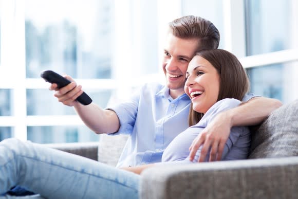 A young couple embraces on the couch and the guy changes the channel with a remote control.