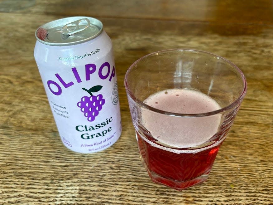 An open can of classic grape Olipop next to a small, clear glass with red liquid inside. Both are sitting on a wooden table.