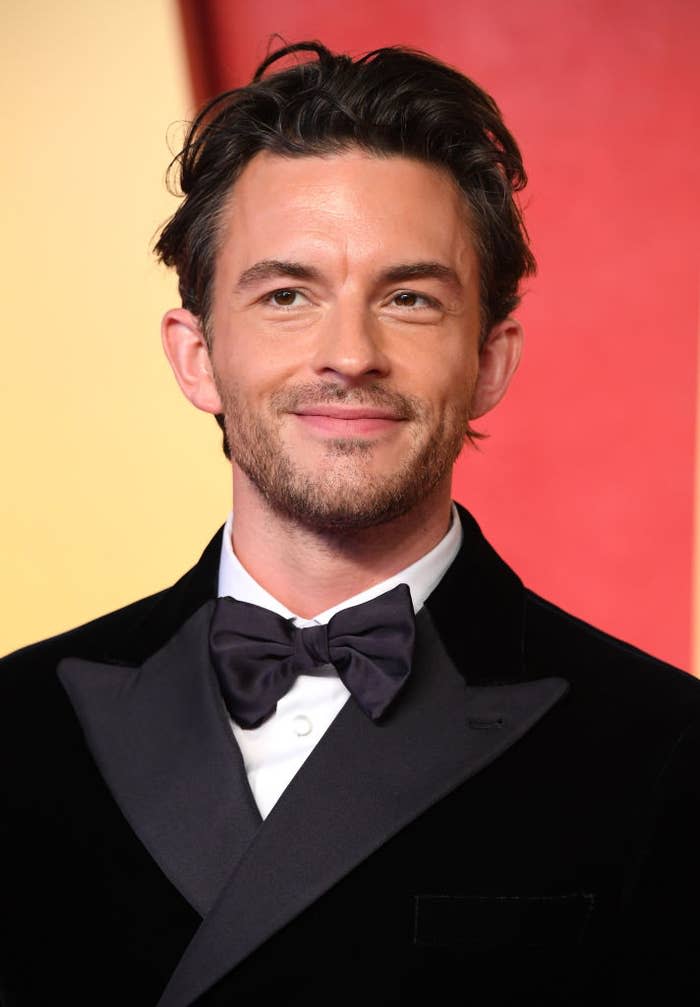 Jonathan Bailey on the red carpet, wearing a classic black tuxedo with a bow tie, smiling for the camera