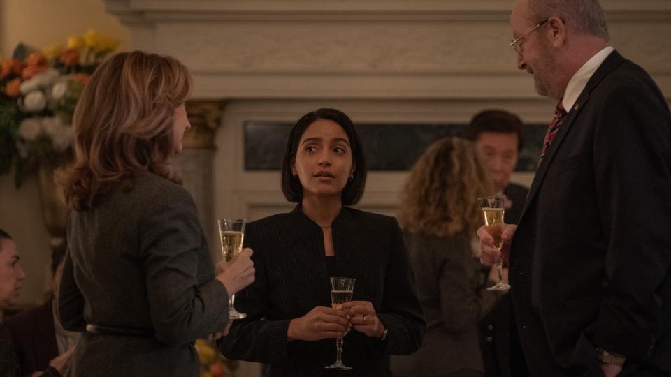 Three well-dressed people are standing together, each holding a glass of champagne. The focus is on the woman standing in the middle.