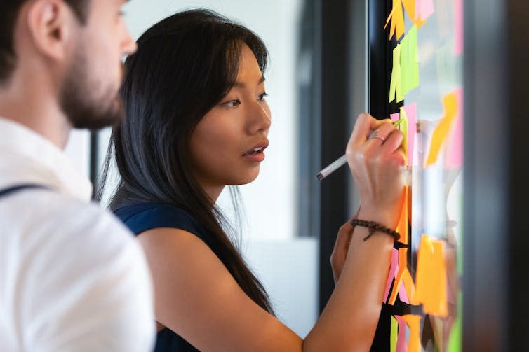 An asian woman writes on post-it-notes stuck on the wall. She wears business attire.