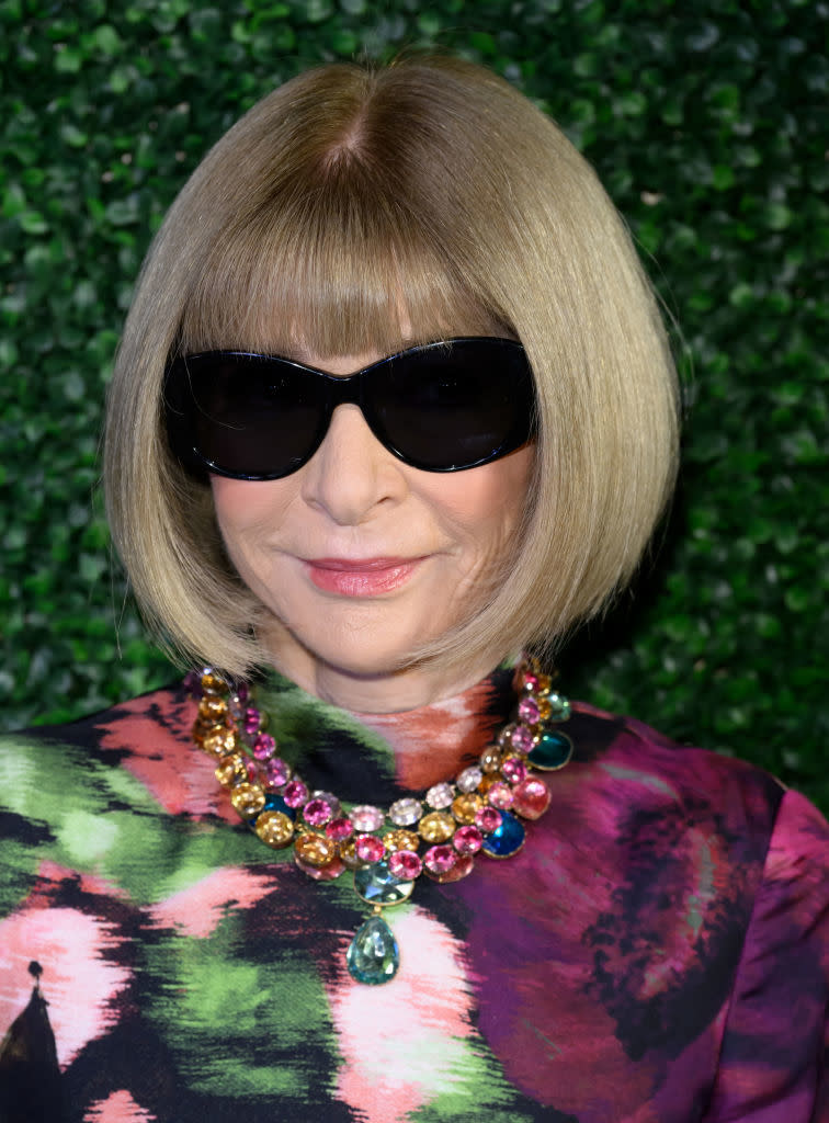 Anna Wintour is pictured wearing large black sunglasses, a colorful patterned top, and a statement necklace adorned with multi-colored gems