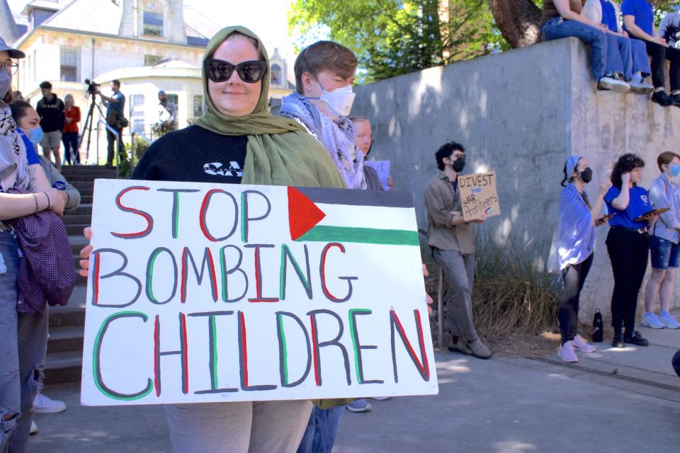 A person wearing sunglasses and a green scarf around their head holds a sign that says "stop bombing children" and a Palestinian flag. 