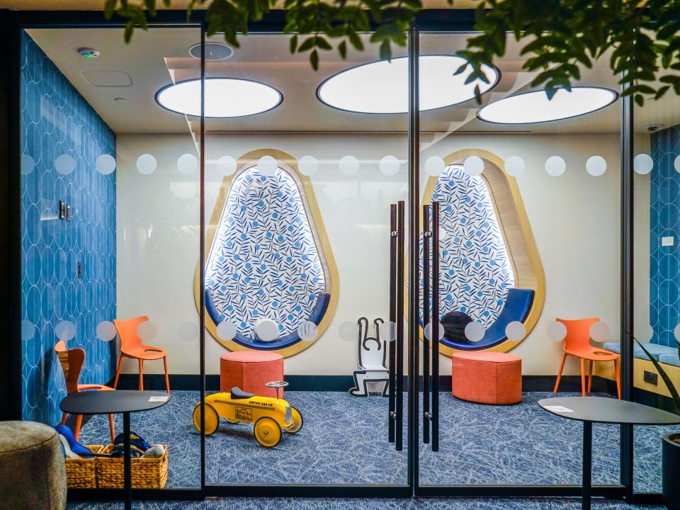 A kids play area with colorful toys and nooks behind glass doors with blue walls and carpets and foliage in hanging over the top of the image