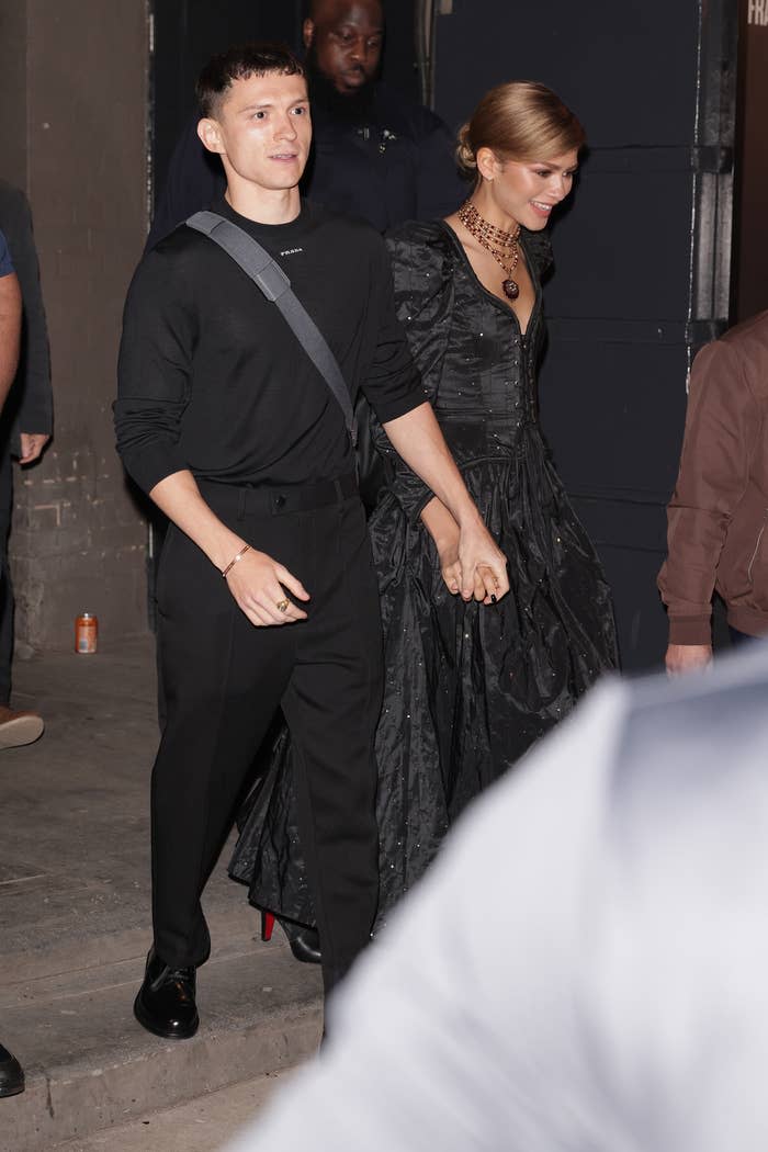 Tom Holland and Zendaya hold hands, with Tom in a black outfit and Zendaya in an elegant black gown with a statement necklace