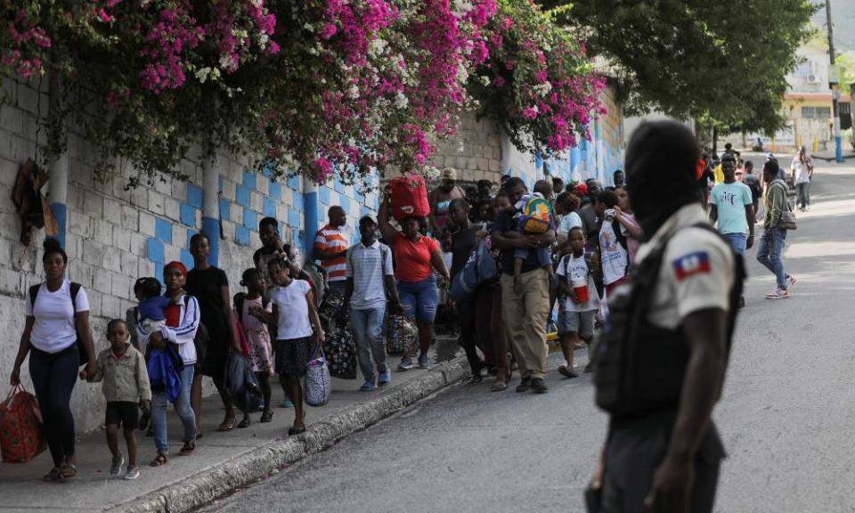 A police officer looks on as dozens of adults and children carrying belongings walk downhill along a road