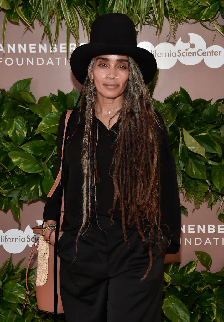 Lisa Bonet in a black outfit and hat, posing in front of a foliage backdrop