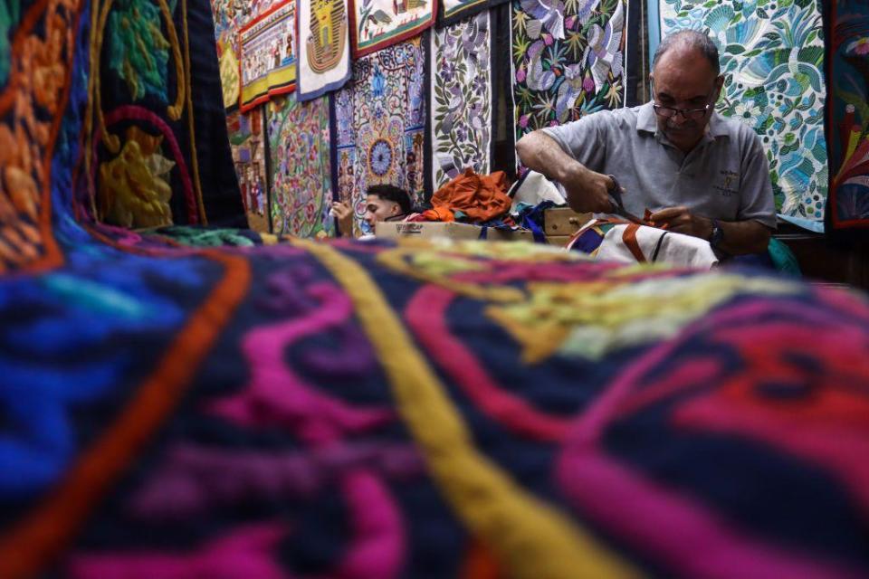 A man sits surrounded by Khayamiya - a decorative Egyptian textile that dates back to Ancient Egypt. He is working on one of the pieces. 