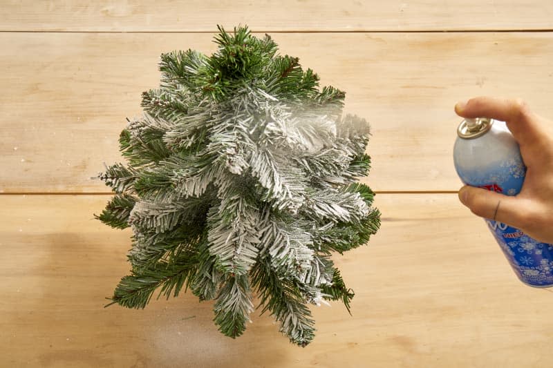 Overhead shot of a small christmas tree being sprayed with fake snow on a light wood surface.